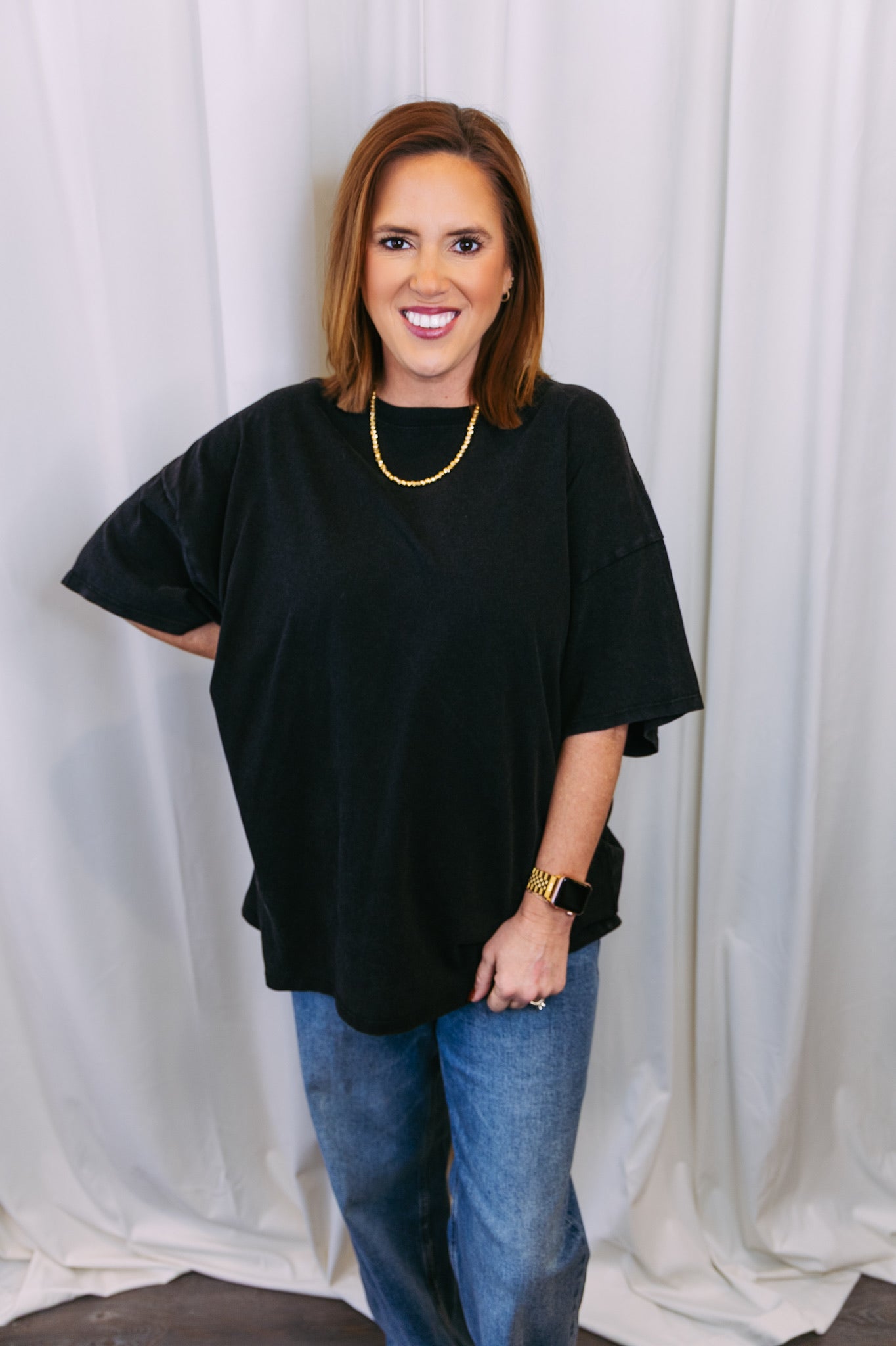 Woman wearing a black top and blue jeans against a white curtain background