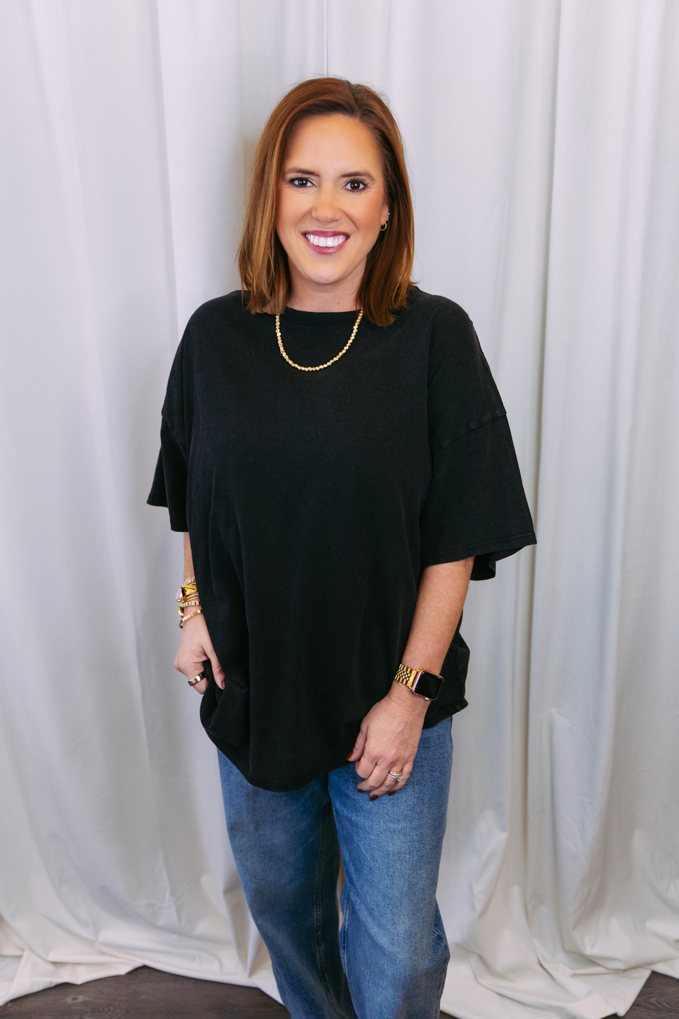 Woman wearing a black top and blue jeans against a white curtain background