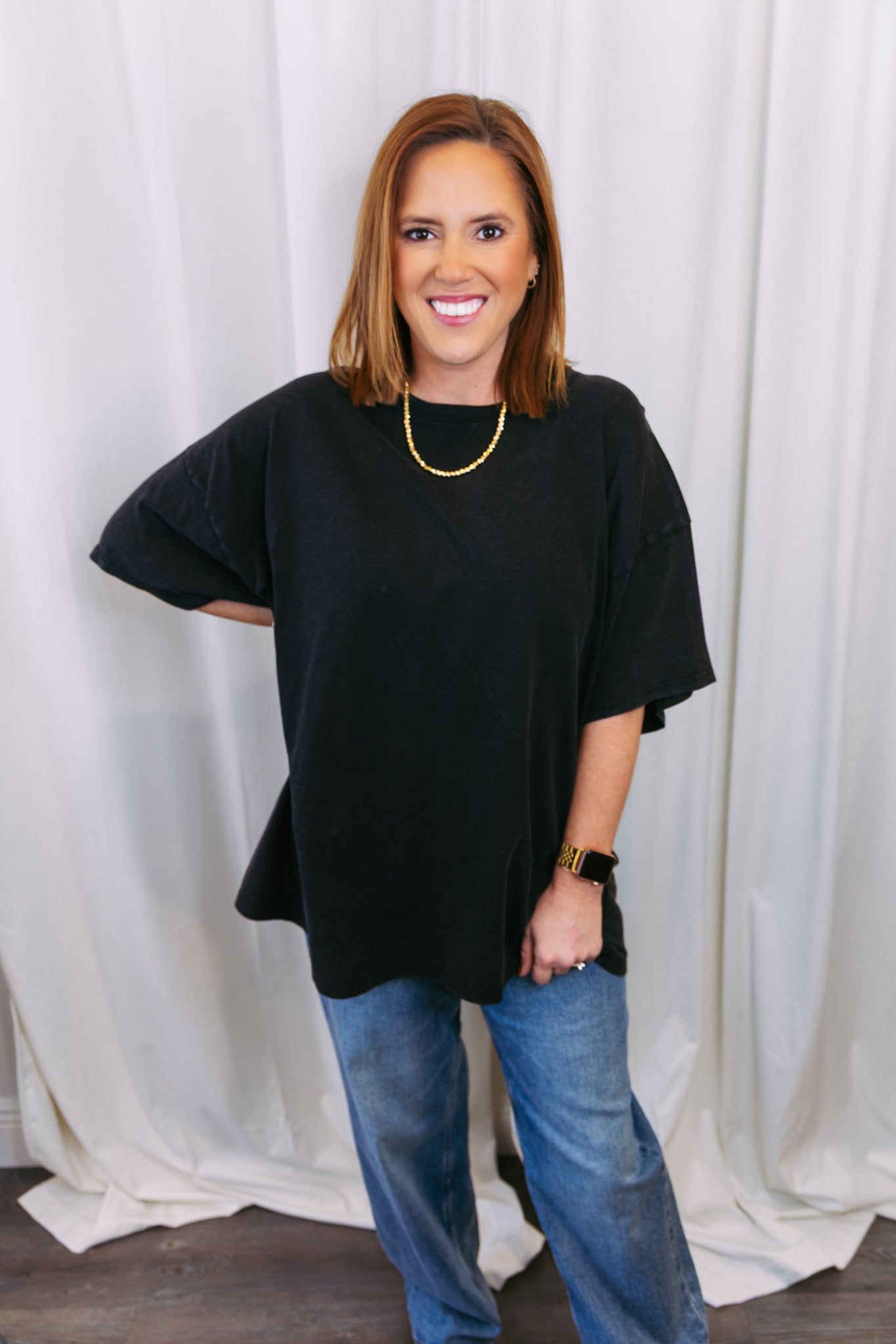 Woman wearing a black oversized top and blue jeans against a white curtain background