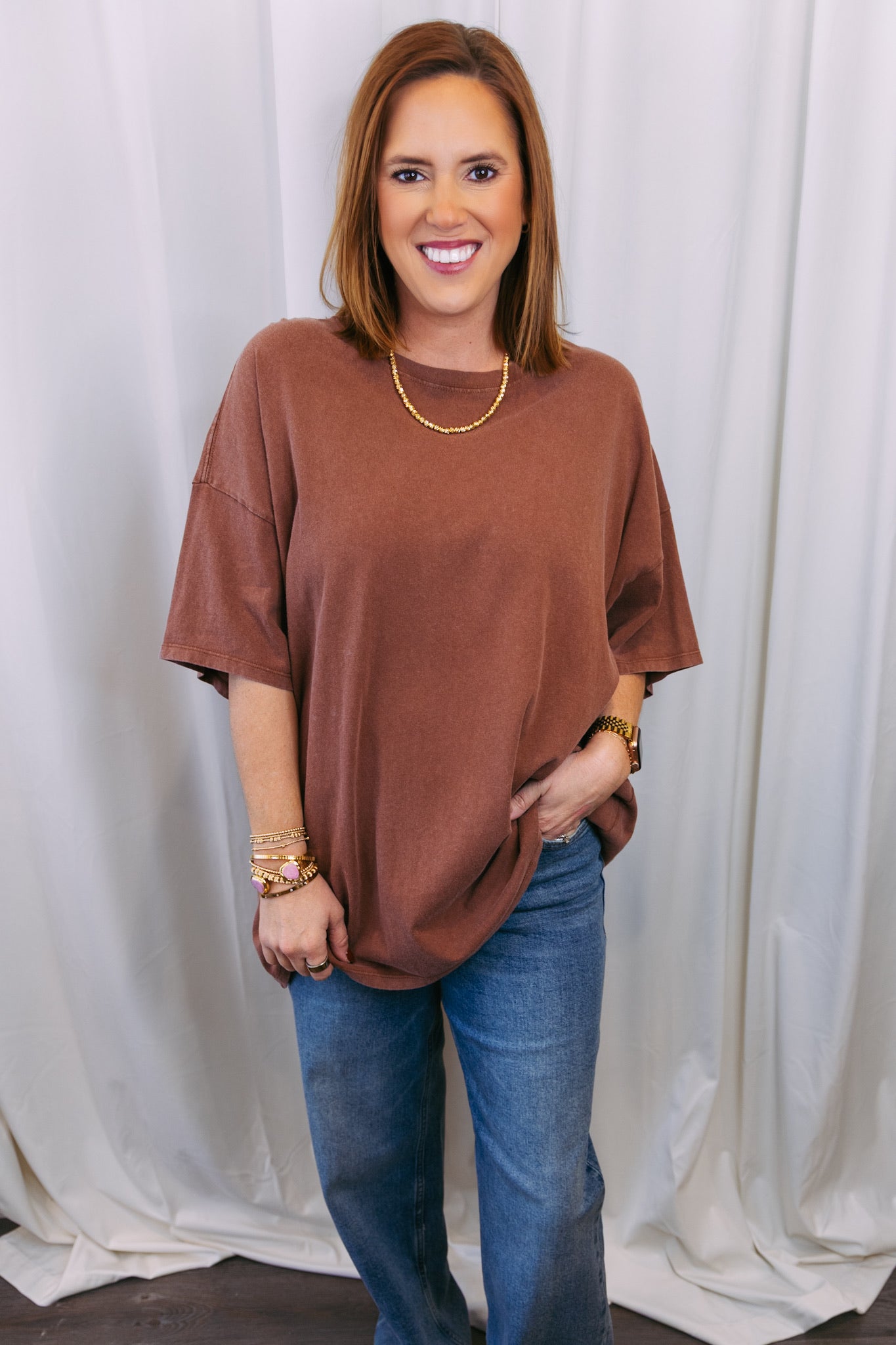 Woman wearing a brown oversized shirt and blue jeans against a white curtain background
