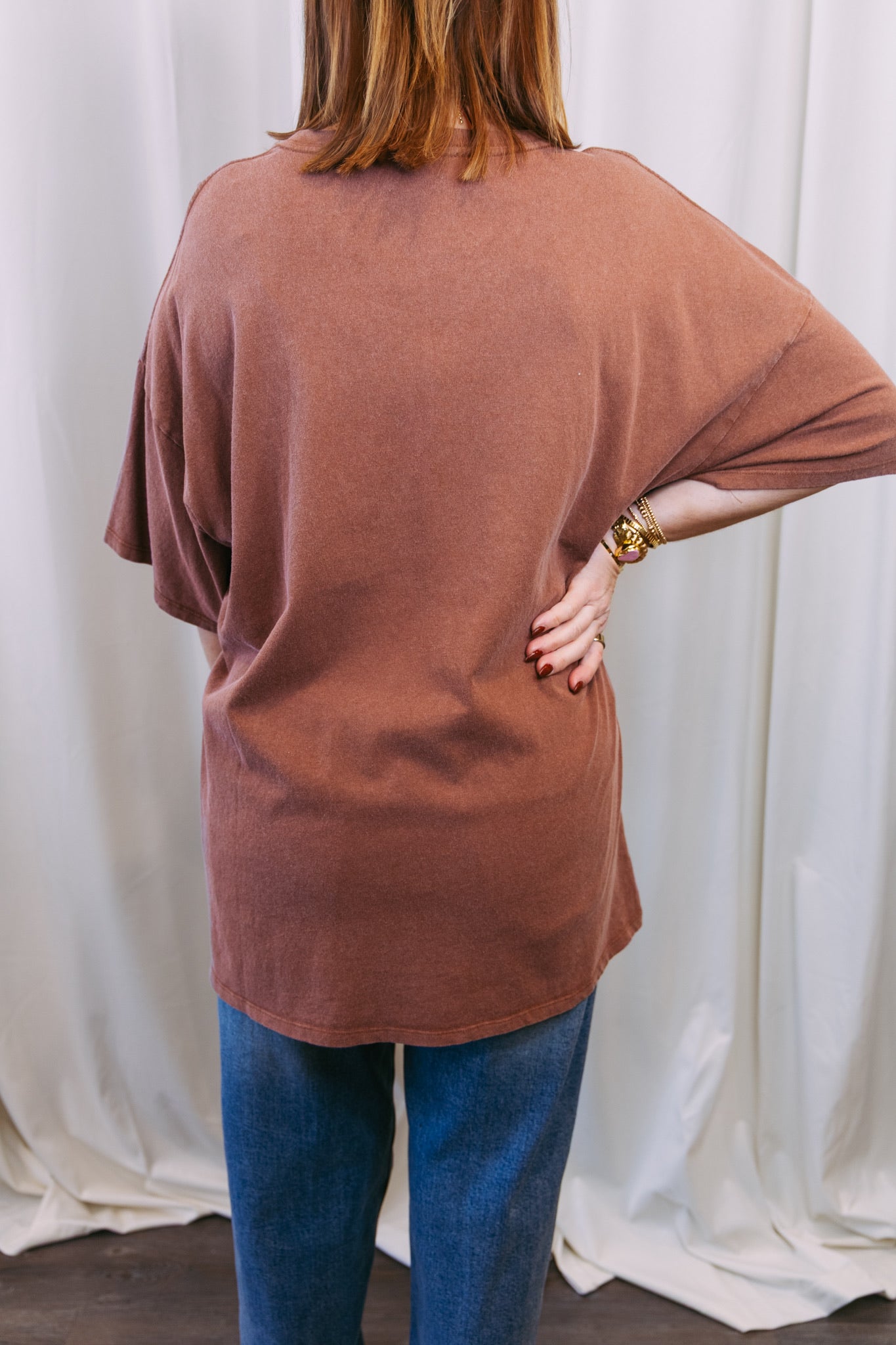 Person wearing a brown oversized shirt and blue jeans against a white curtain background