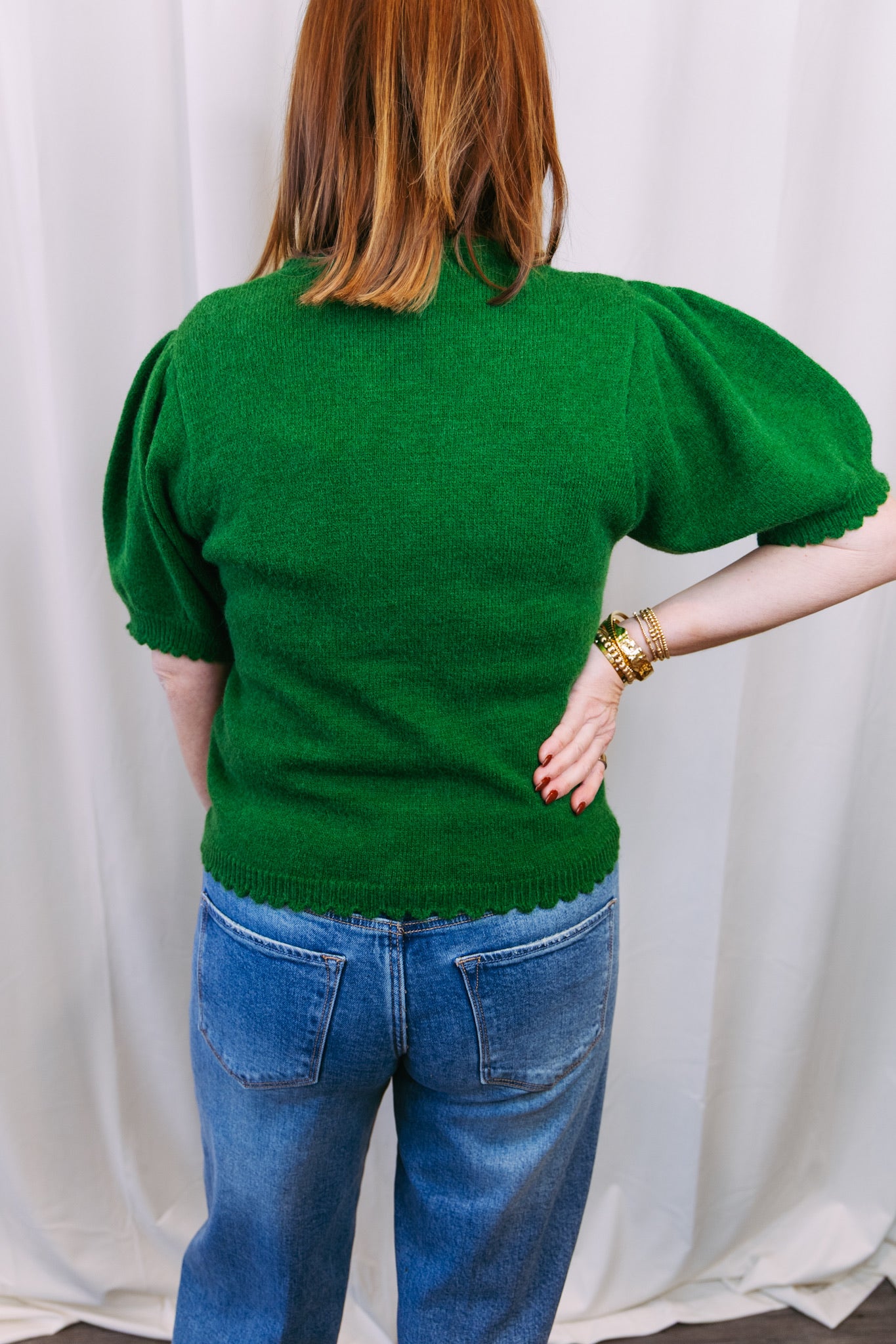 Person wearing a green sweater and blue jeans against a white curtain background