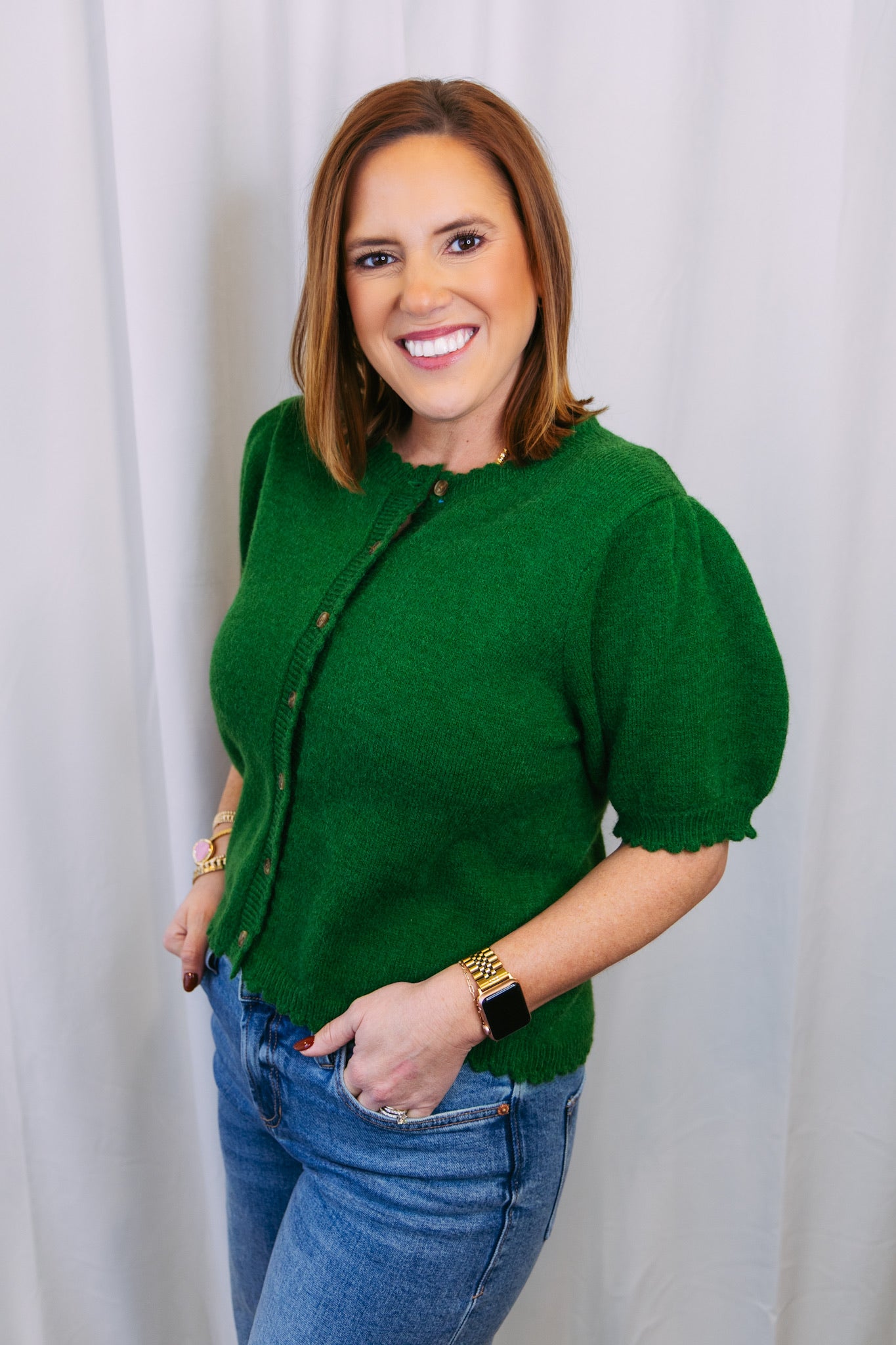 Woman wearing a green sweater and blue jeans against a white curtain background
