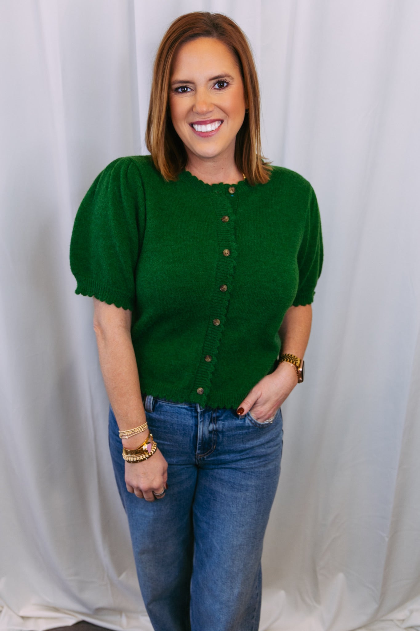 Woman wearing a green sweater with puff sleeves and blue jeans against a white curtain background