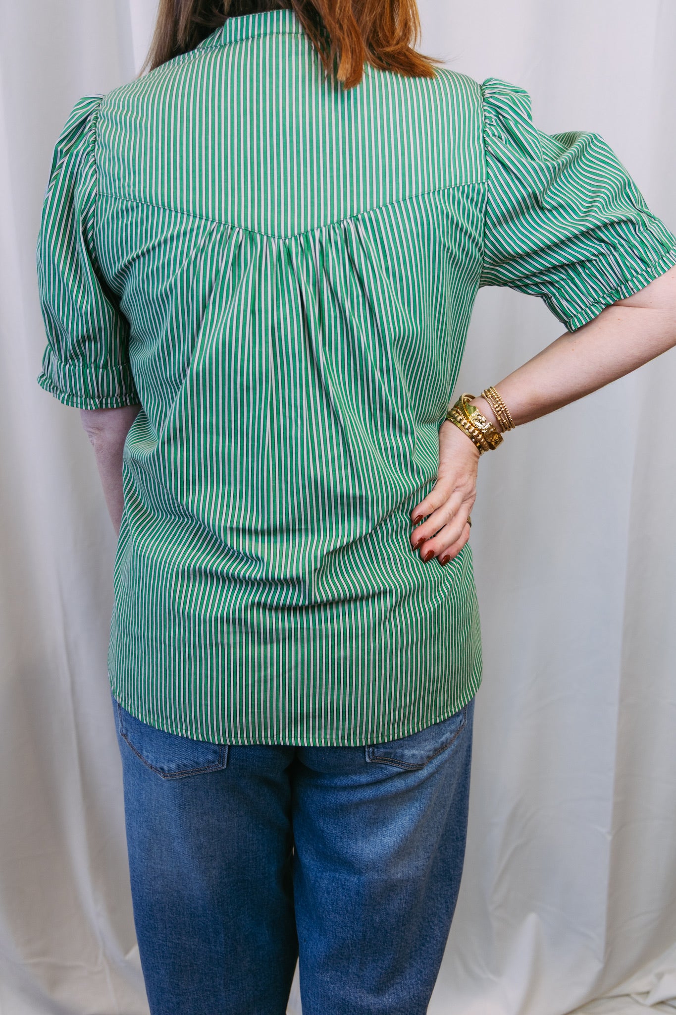 Person wearing a green striped shirt and blue jeans against a white curtain background