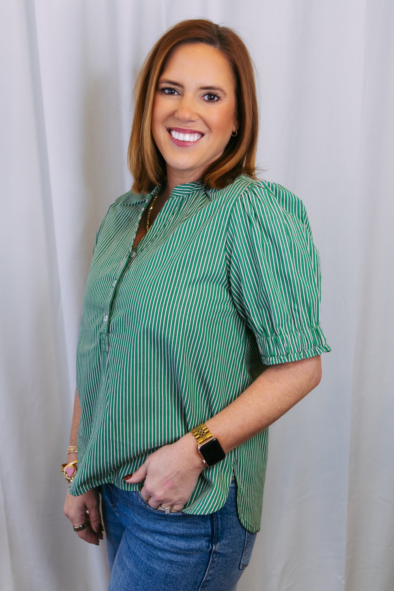 Woman wearing a green striped shirt and blue jeans against a white curtain background