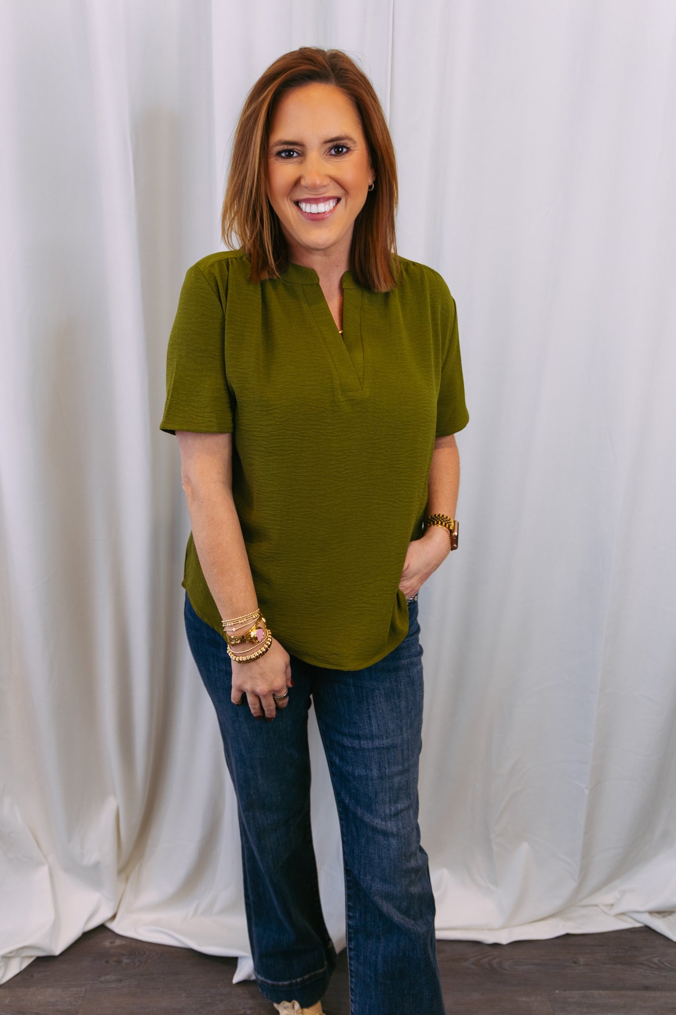 Woman wearing an olive green top and blue jeans standing against a white curtain.