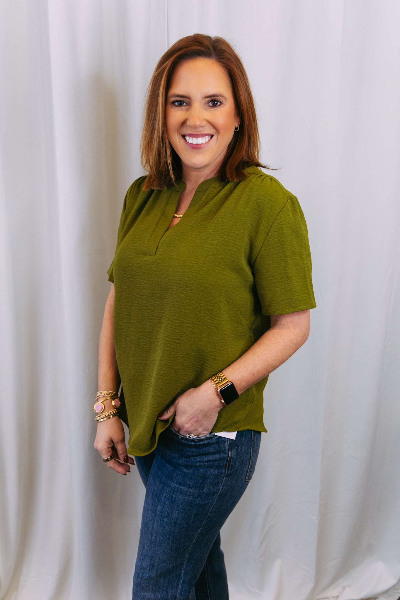 Woman wearing a green top and blue jeans against a white curtain background
