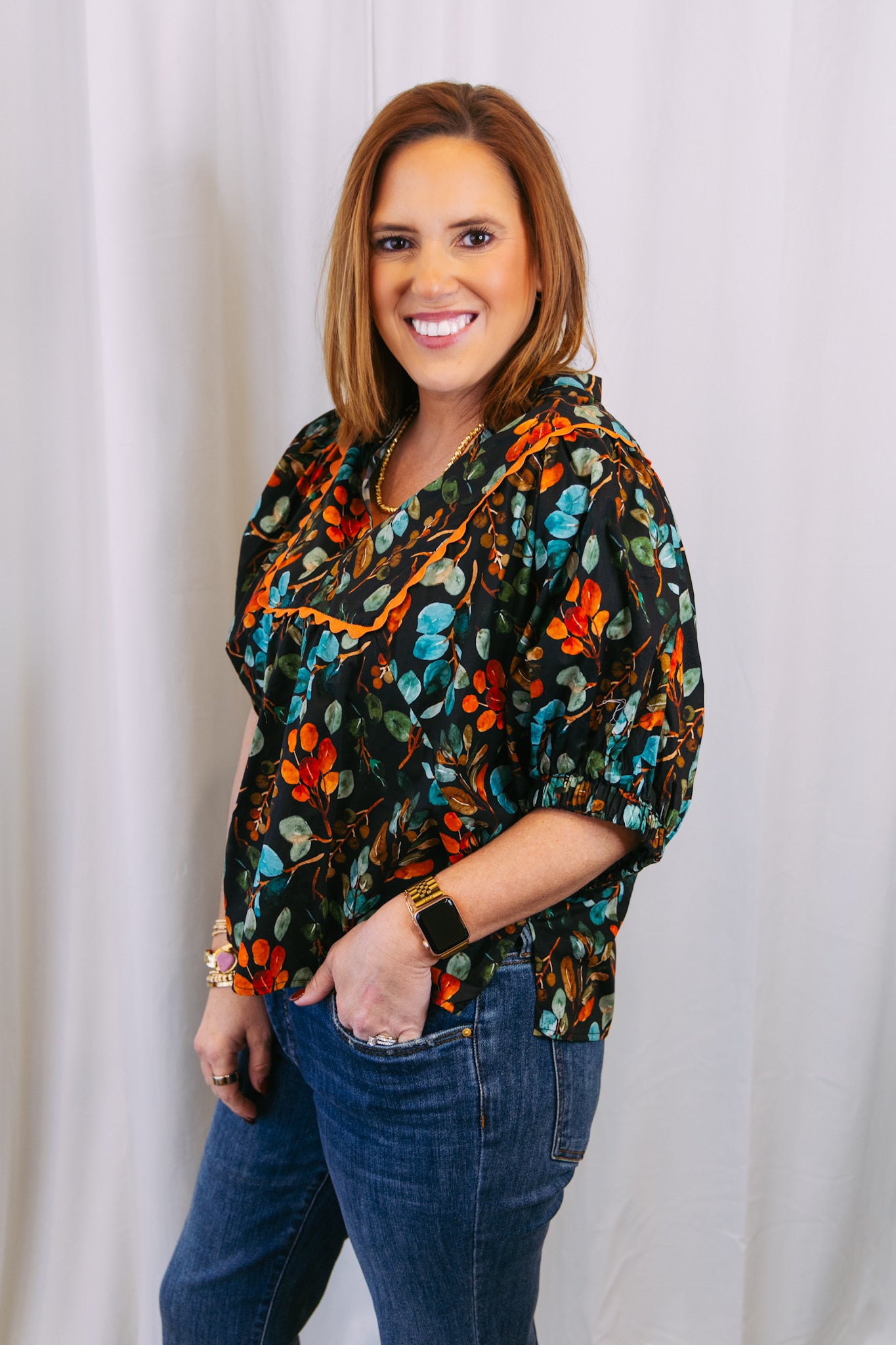 Woman wearing a colorful floral blouse and jeans against a white curtain background