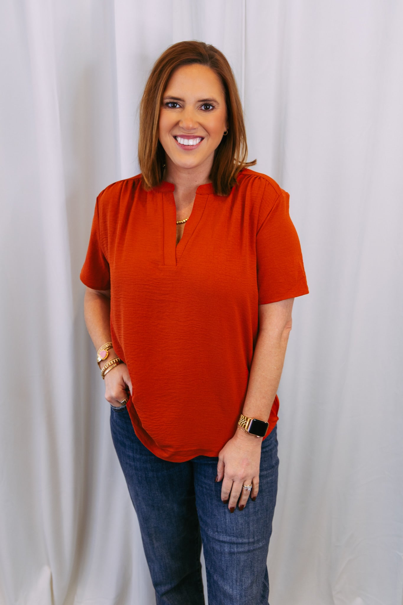 Woman wearing a rust-colored top and blue jeans against a white curtain background