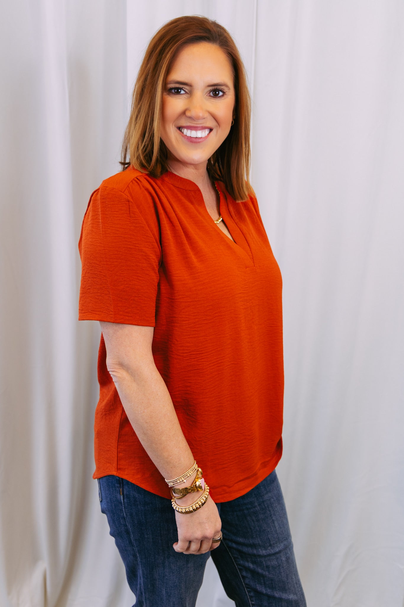 Woman wearing an rust top and blue jeans against a white curtain background