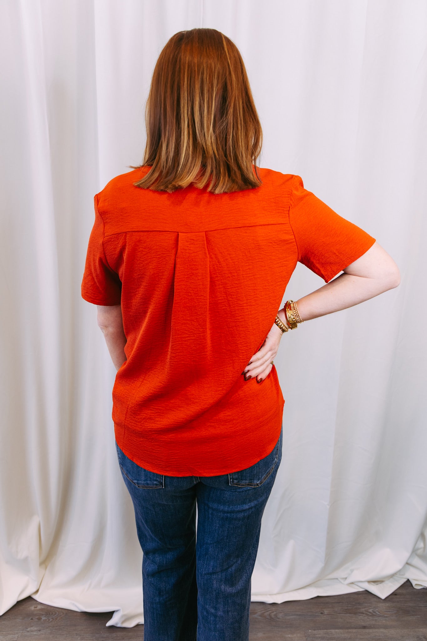 Person wearing an rust top and blue jeans standing against a white curtain.