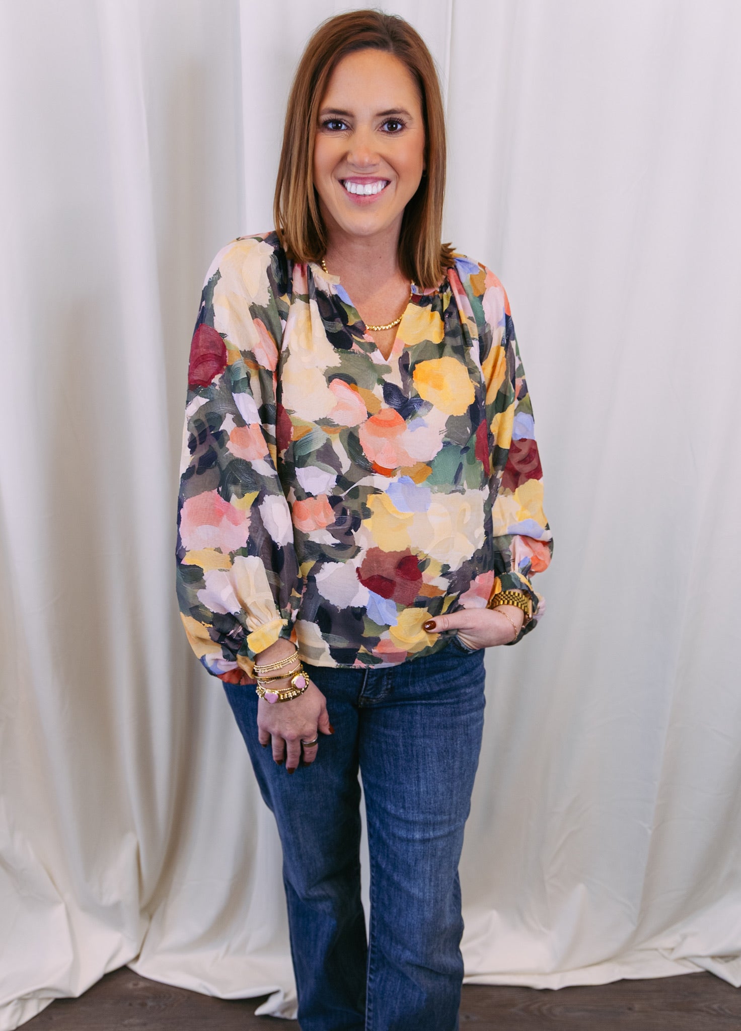 Woman wearing a colorful floral sweater and blue jeans against a white curtain background