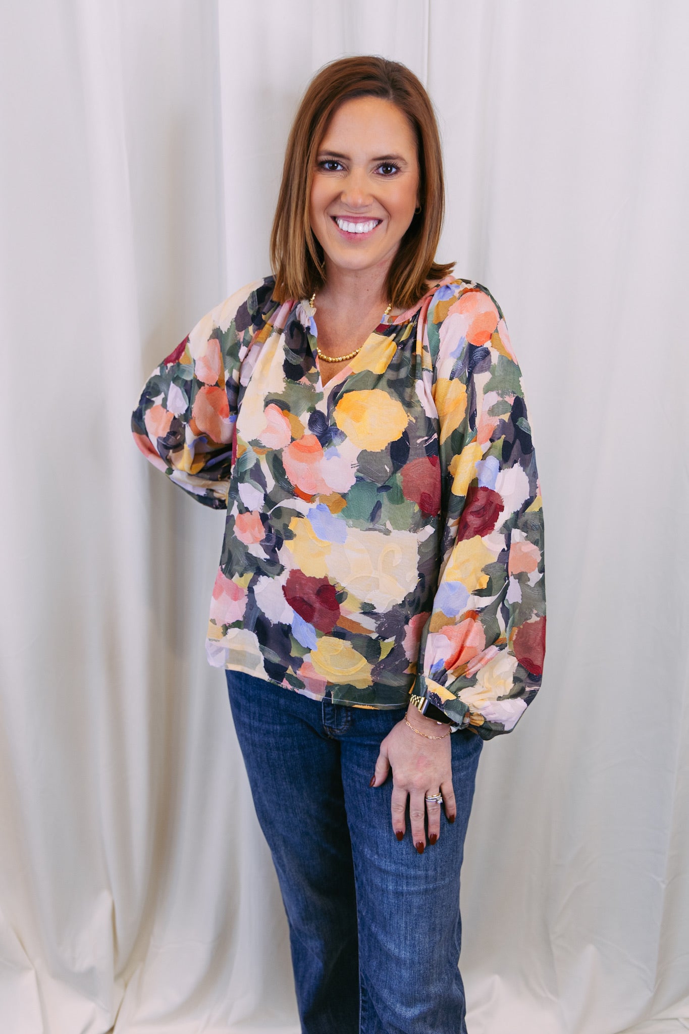 Woman wearing a colorful floral blouse and jeans against a white curtain background