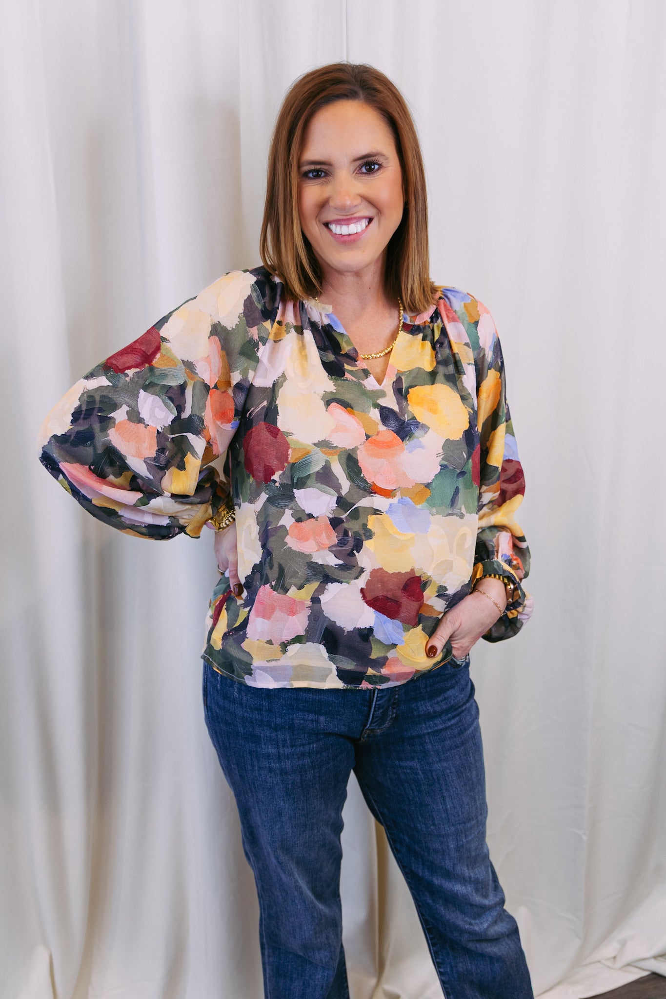 Woman wearing a colorful floral blouse and blue jeans against a white curtain background
