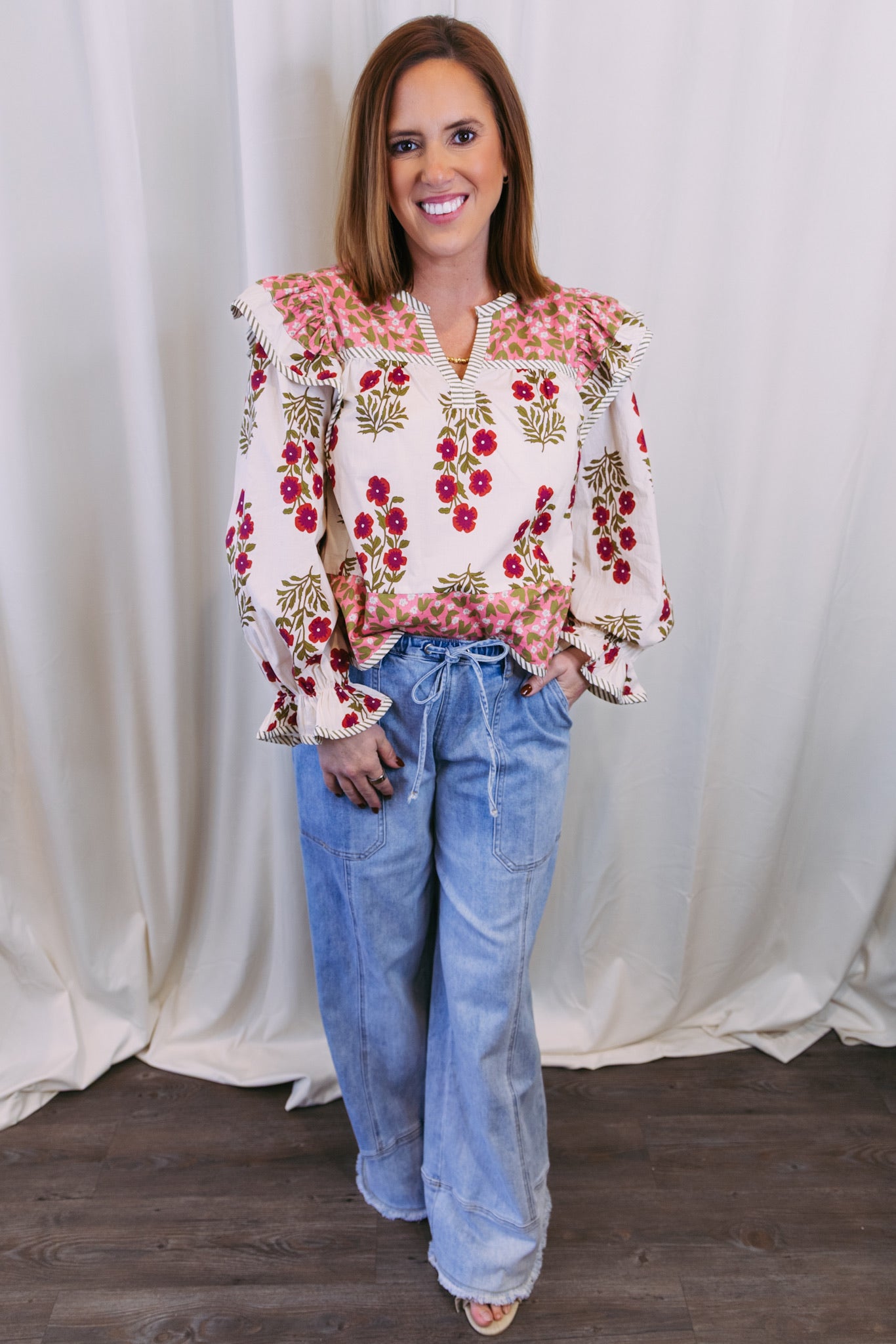 Woman wearing a floral blouse and jeans against a white curtain background