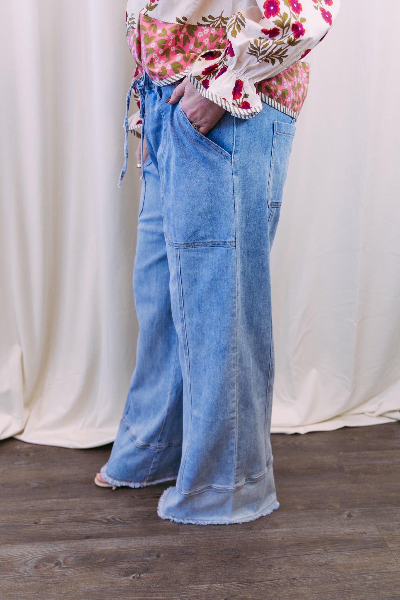 Person wearing blue jeans and a floral top against a white curtain background