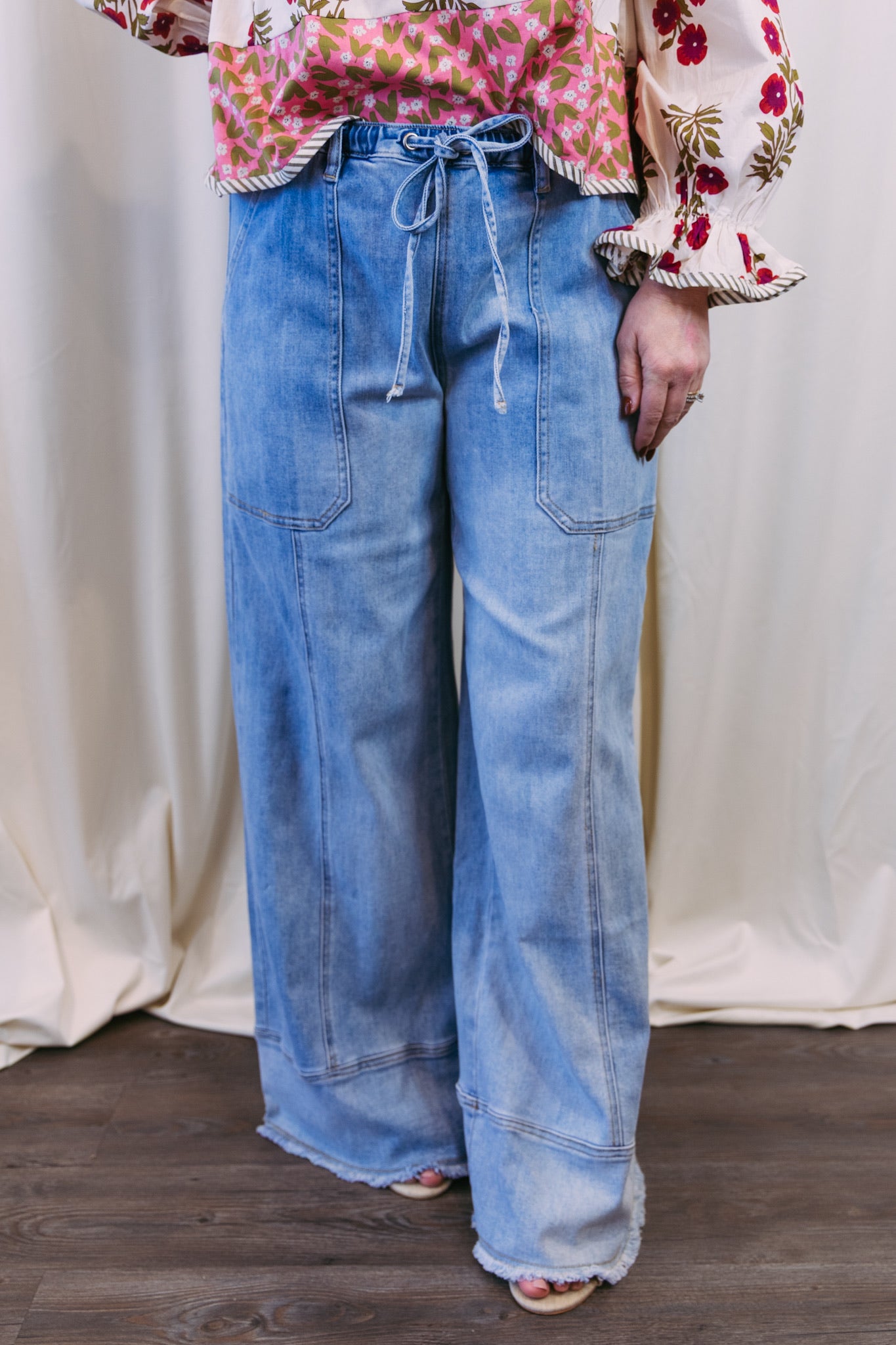 Person wearing a floral blouse and blue jeans against a white curtain background