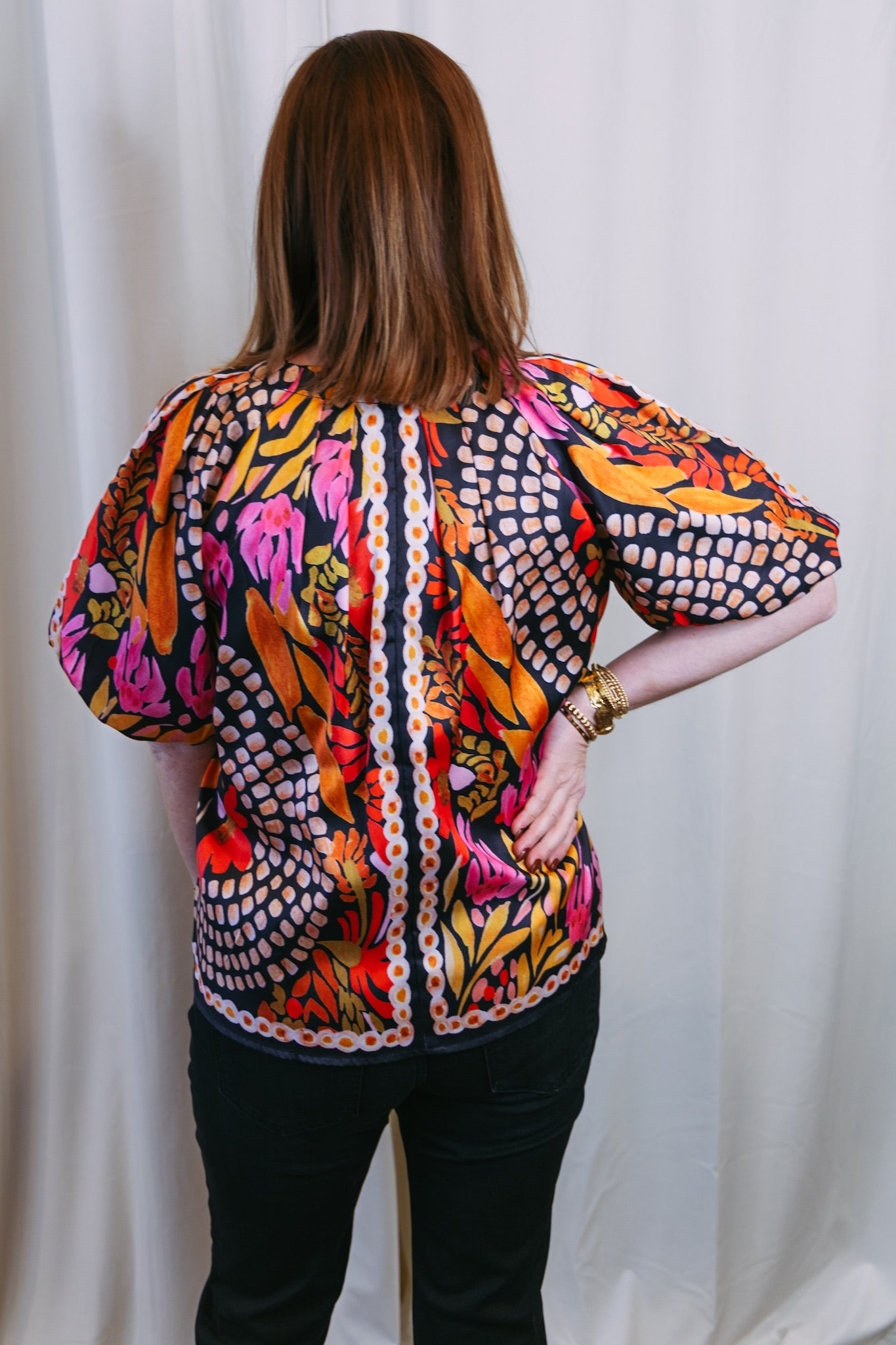 Person wearing a colorful patterned blouse with black pants against a white curtain background