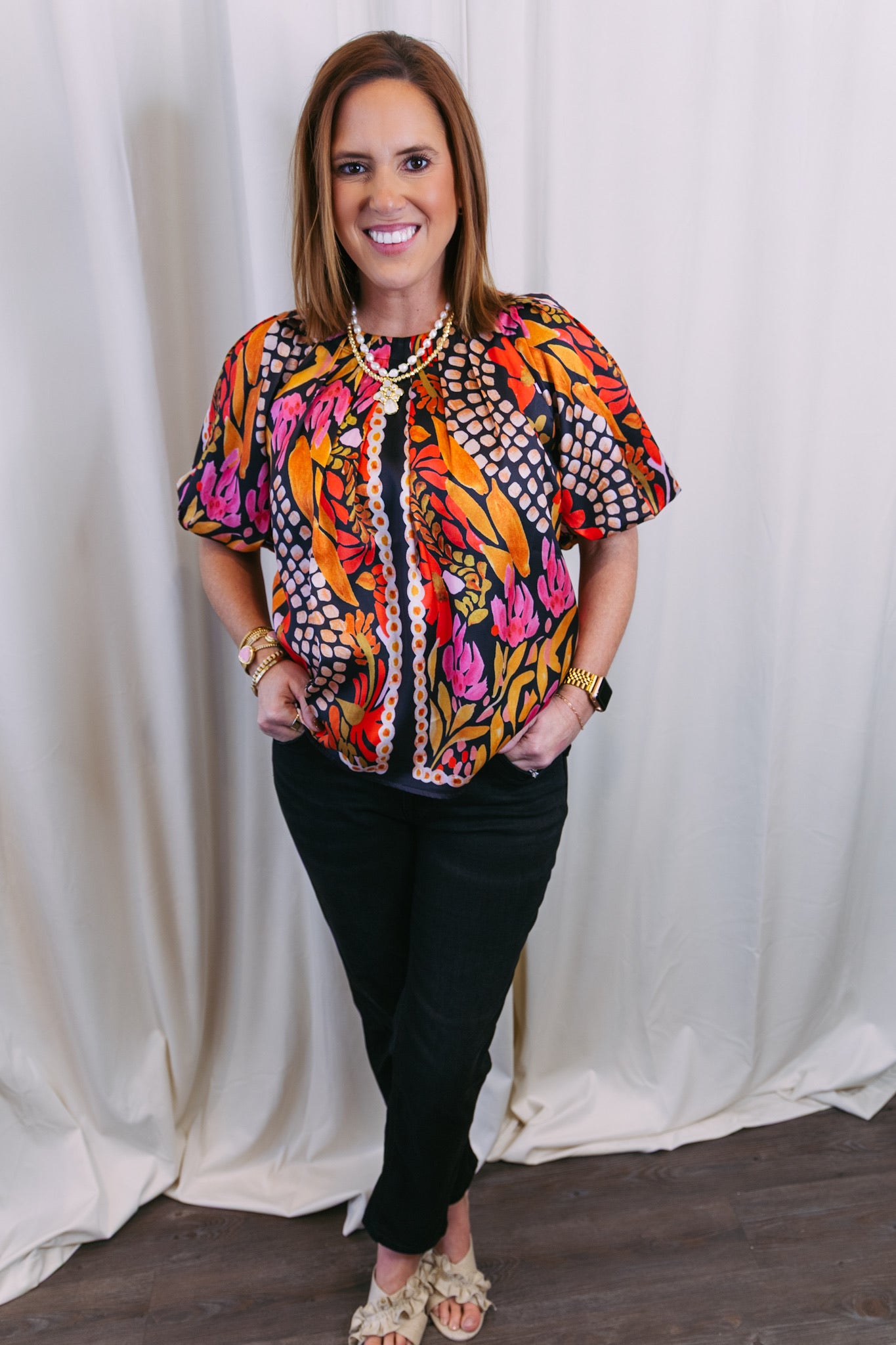 Woman wearing a colorful patterned blouse and black pants against a white curtain background