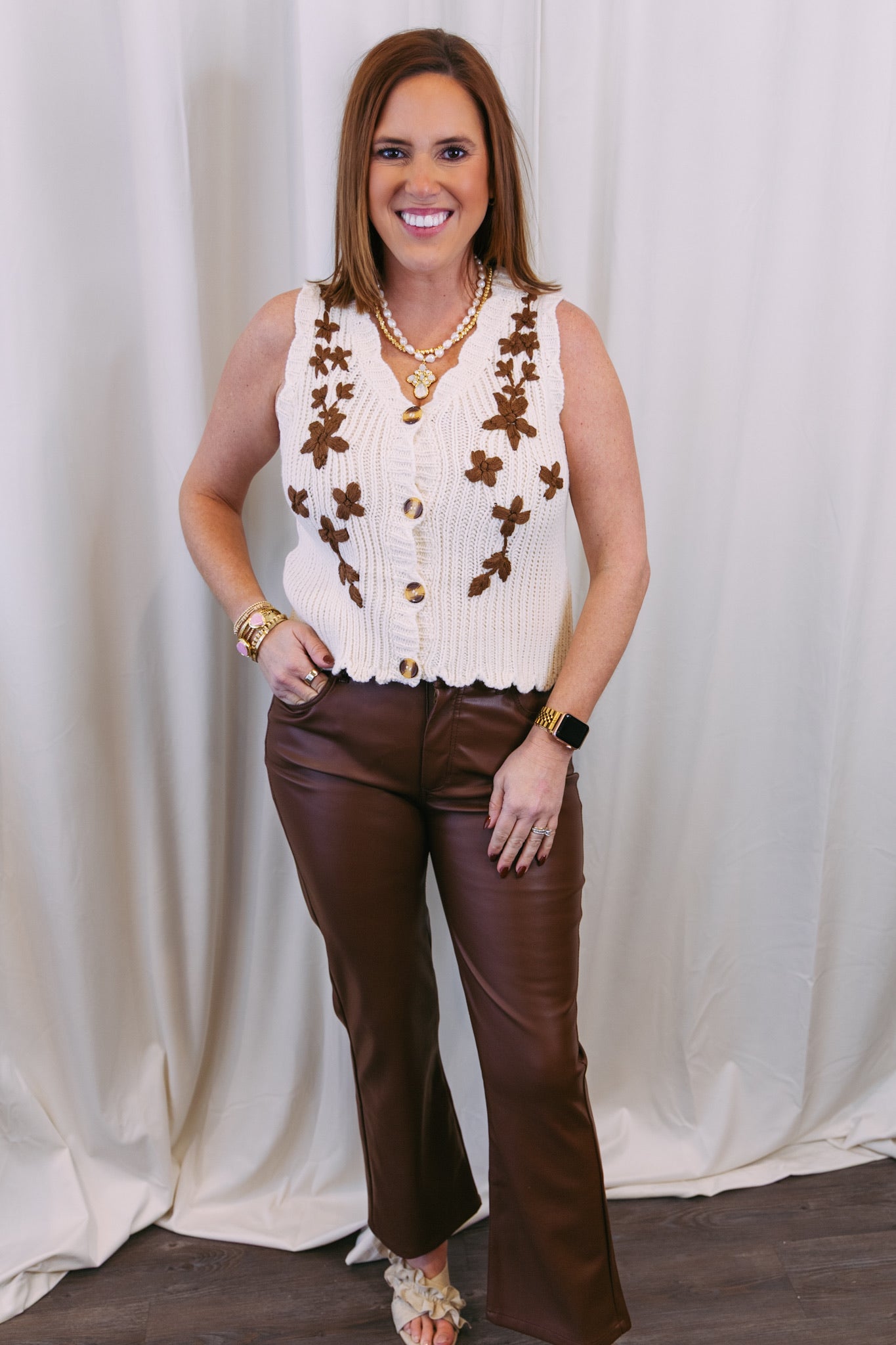 Woman wearing a white sleeveless floral vest and brown bell-bottom pants against a white curtain.