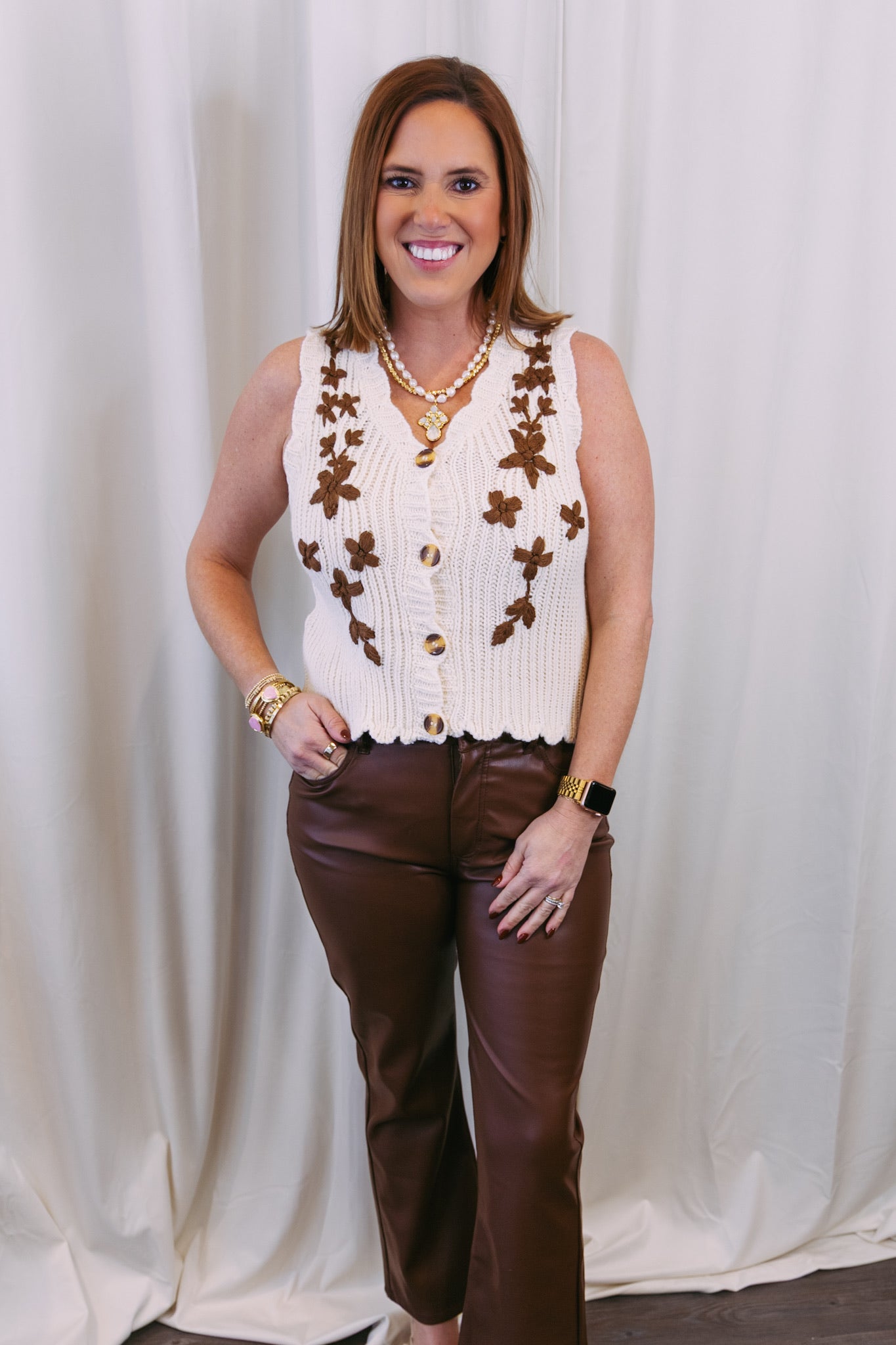 Woman wearing a cream-colored vest with brown floral patterns and brown pants against a white curtain background.
