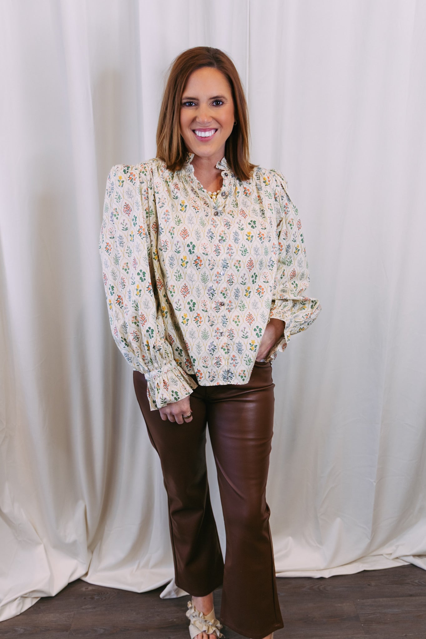 Woman wearing a floral blouse and brown pants against a white curtain background