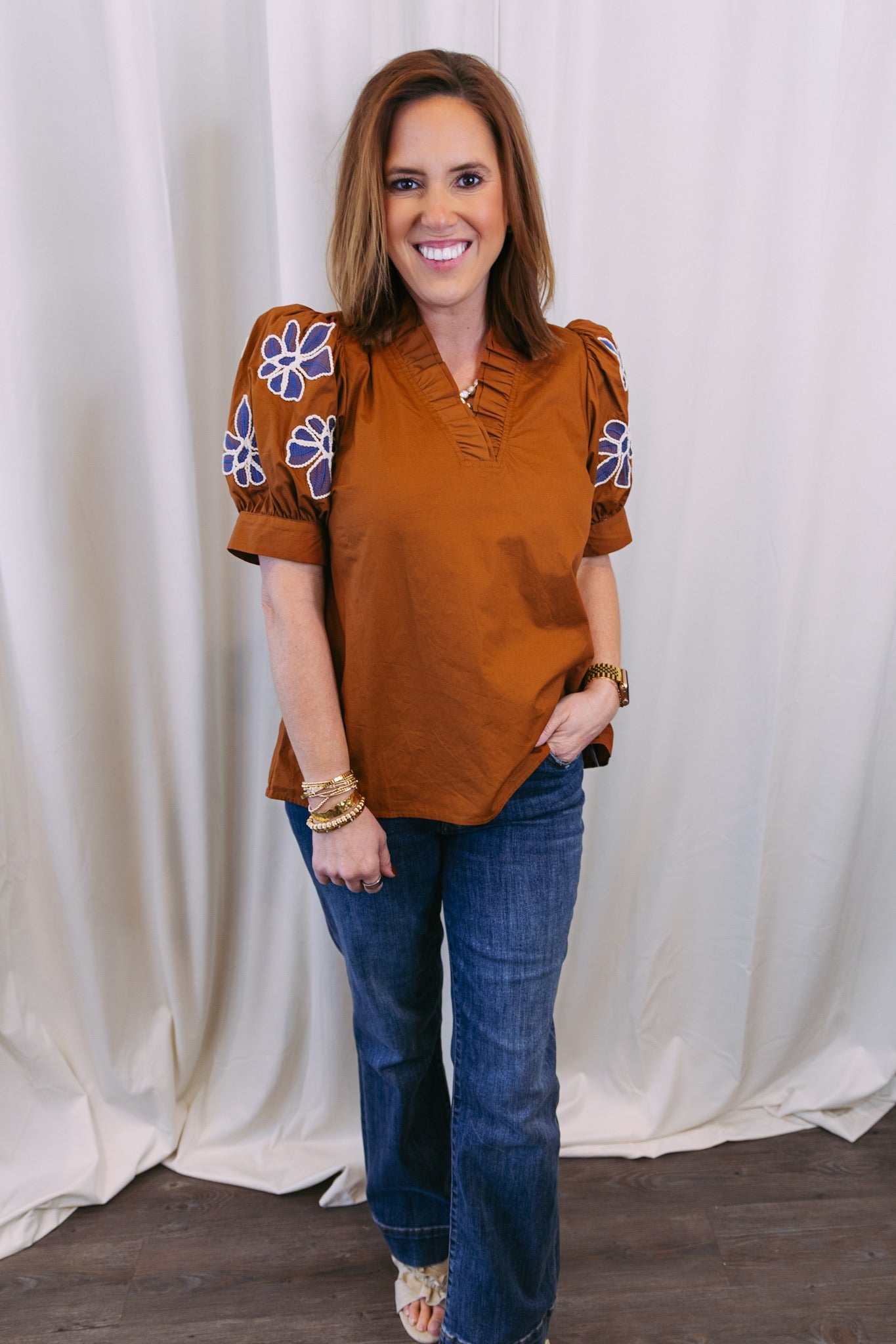 Woman wearing a brown blouse with floral sleeves and blue jeans against a white curtain background