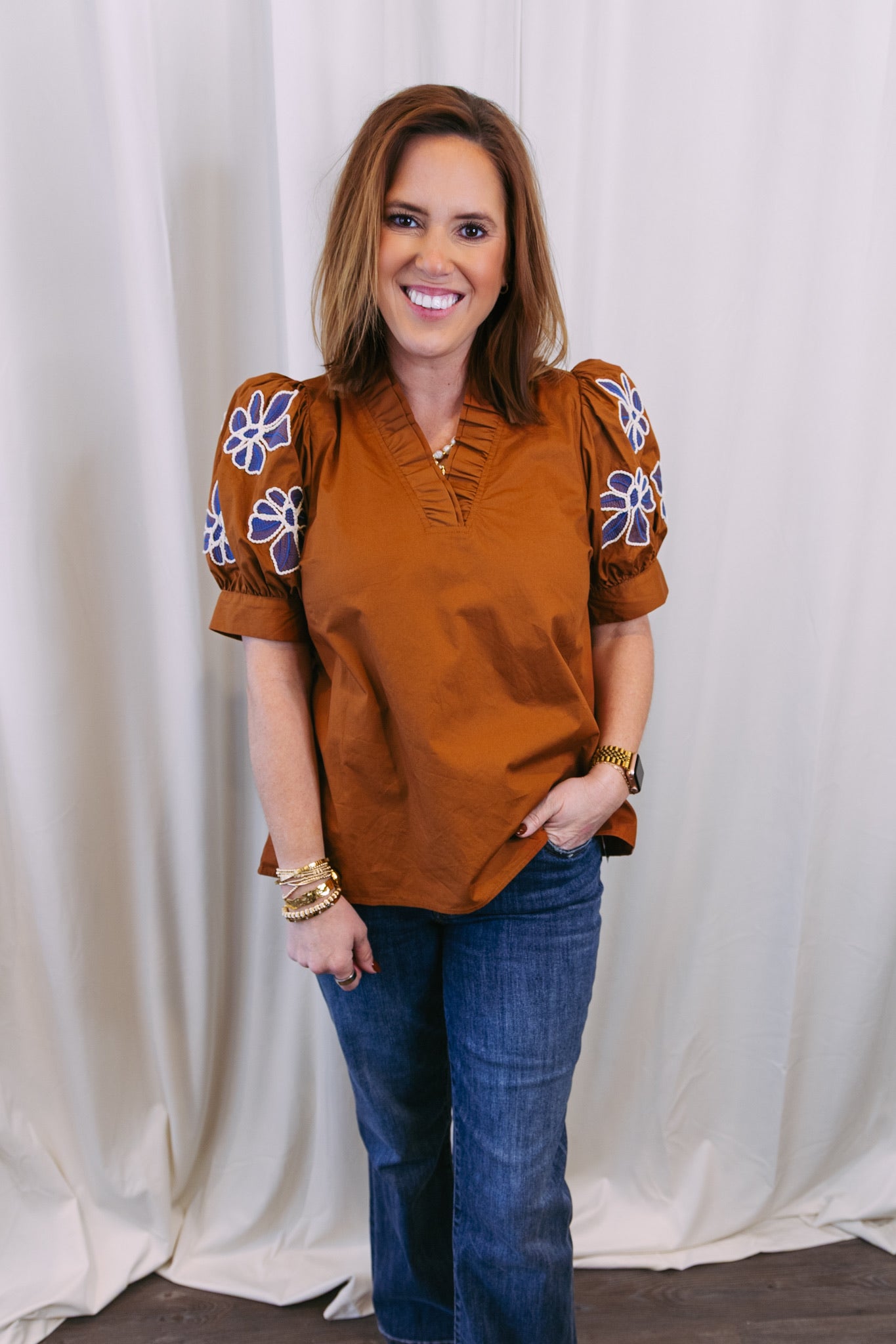 Woman wearing a rust-colored blouse with floral patterns on sleeves against a white curtain background